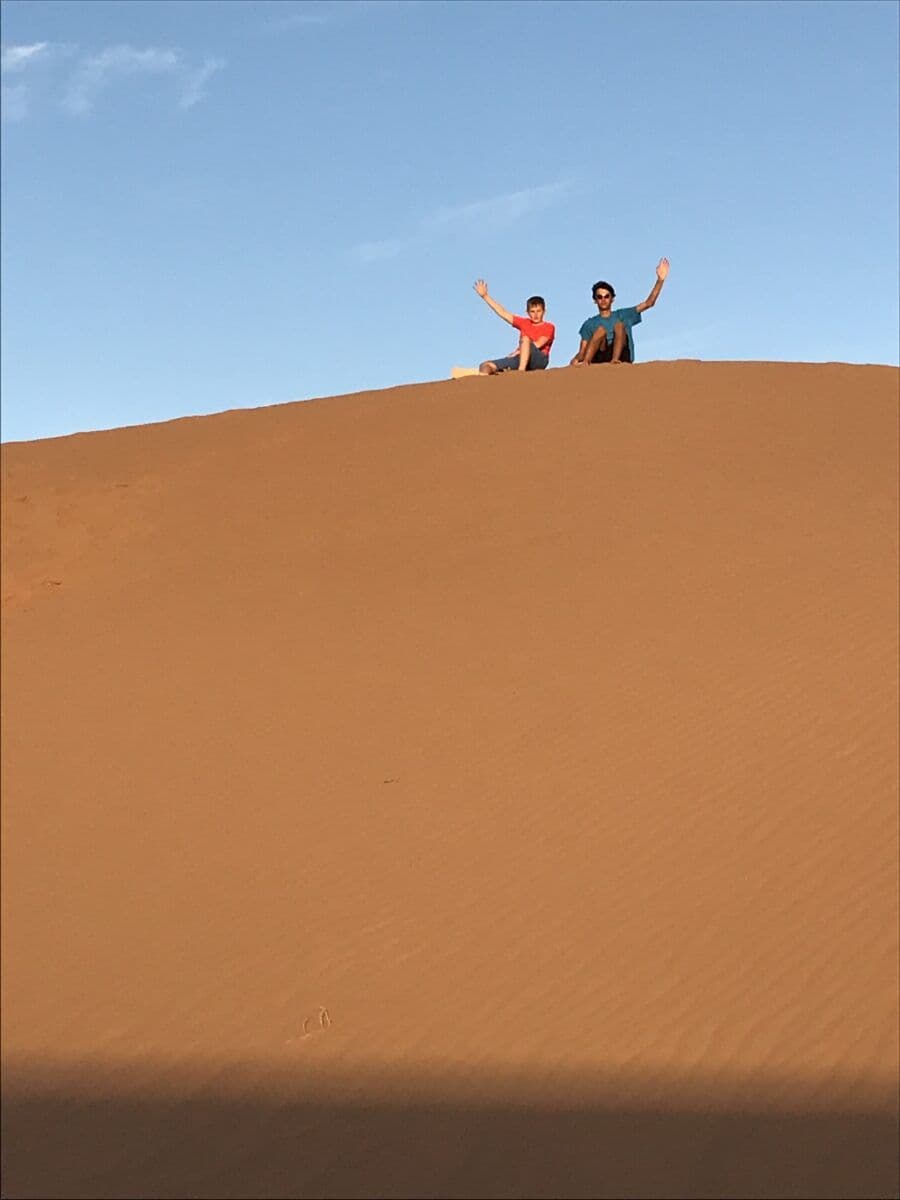 Twee deelnemers zwaaien vanop de top van een zandduin in de Sahara